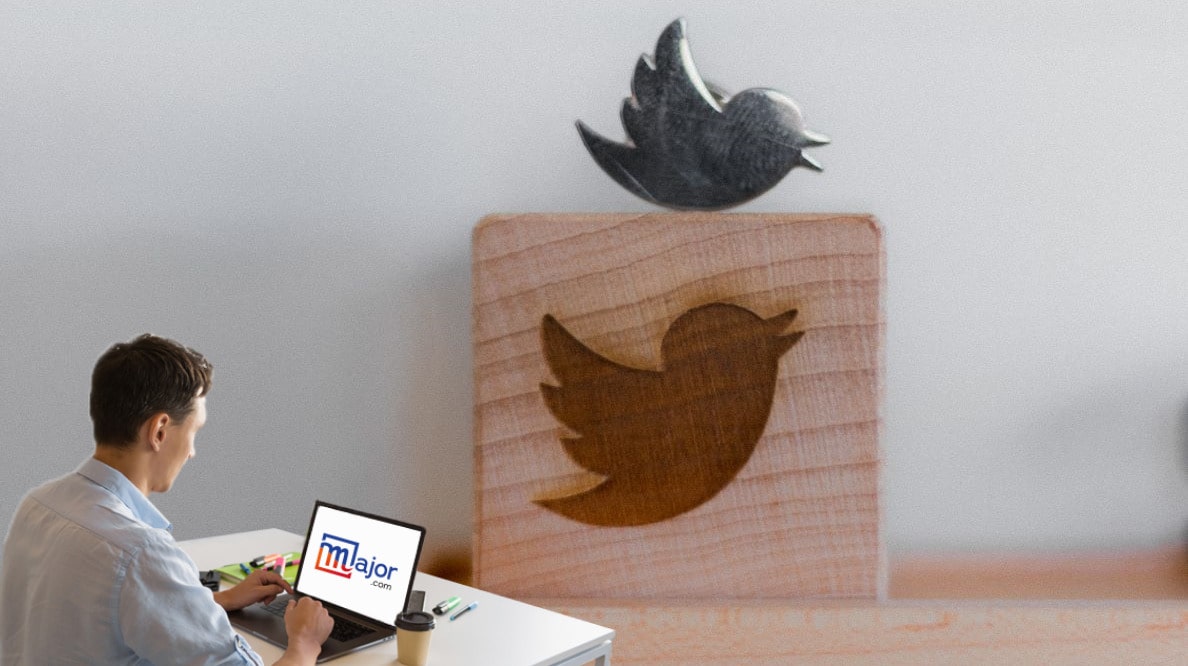 Un homme travaillant sur un ordinateur avec le logo Major.com devant une sculpture en bois représentant le logo de Twitter, un oiseau