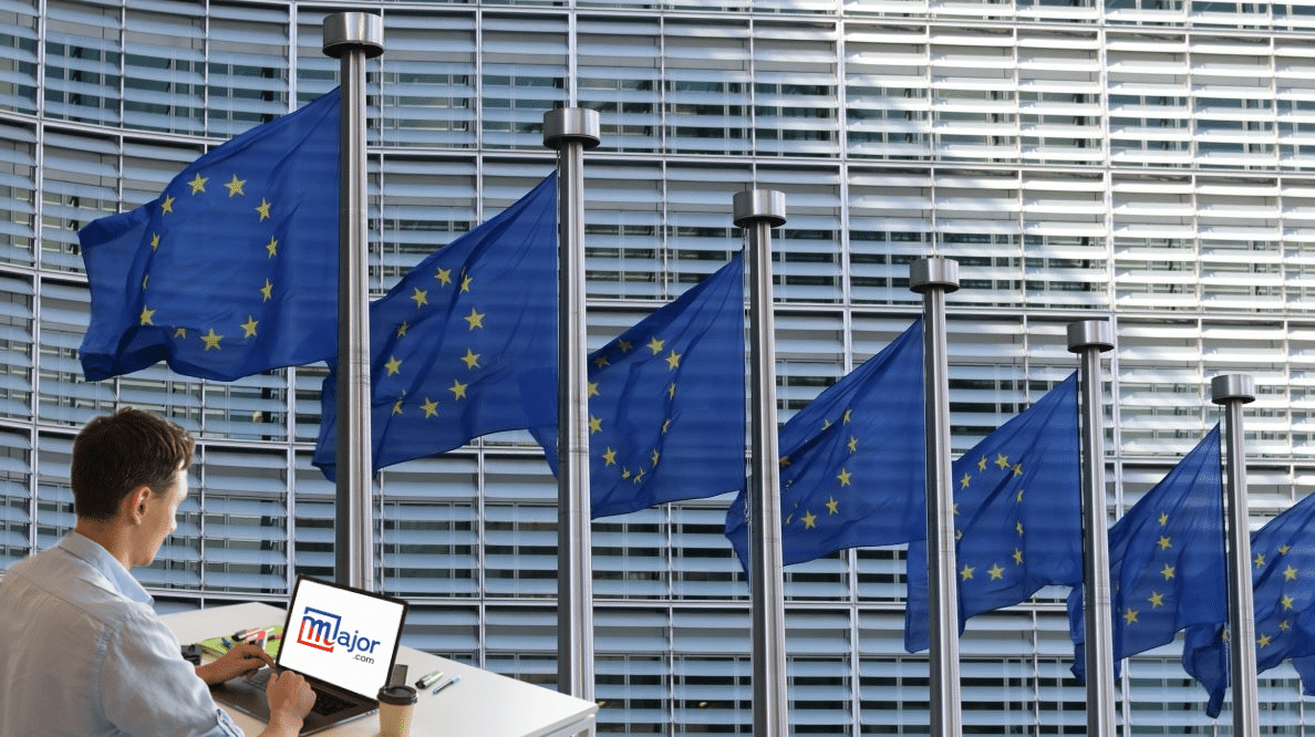 Un homme est à son bureau devant son ordinateur qui porte le logo du média Major. Derrière, des drapeaux européens flottent