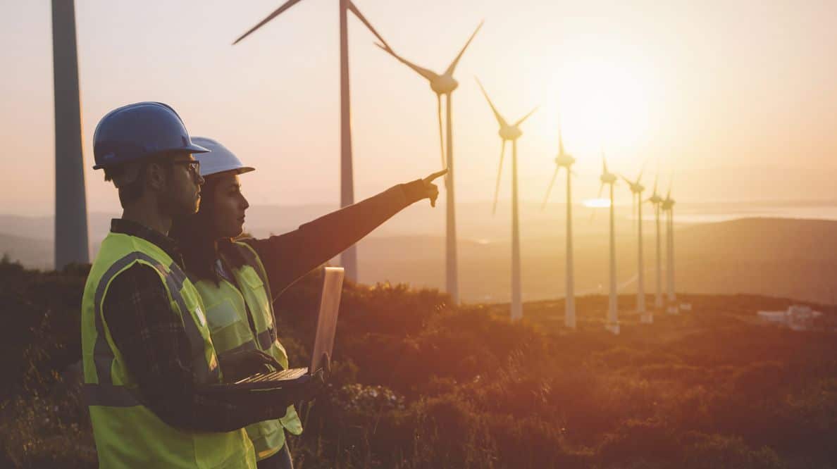 Deux personnes portant un gilet jaune et un casque de protection discutant devant des éoliennes au soleil couchant.