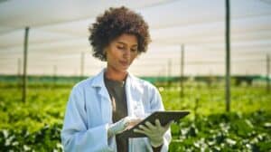 Une femme métisse dans un champ vert portant une blouse blanche et manipulant une tablette électronique